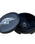 A black round metal tin with its lid leaning against it on a white background. The lid features a graphic of a logo while the inside of the tin is also black with a logo that reads 'SUCK LESS.