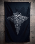 A black flag hanging on a textured concrete wall, featuring a large silver metallic graphic of a winged caduceus skull in the center.