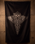 A black flag featuring a detailed silver graphic of a winged caduceus with a skull and crown, hanging against a textured concrete wall.
