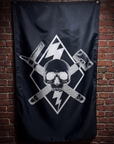 A black flag featuring a white skull and crossed tools graphic, hanging against a red-brown brick wall.
