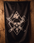 A black flag featuring a white skull and crossed tools graphic, hanging against a rustic wooden plank wall.