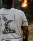 A man viewed from behind wearing a black baseball cap and a light tan t-shirt with a large black graphic print of an angel on the back, set against a blurred outdoor background.