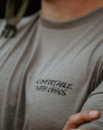 A close-up lifestyle shot of a man wearing a gray crewneck t-shirt with the black printed text 'COMFORTABLE WITH CHAOS' on the left chest. He is also wearing a black tactical headset.