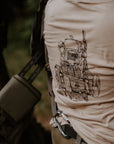 A close-up lifestyle shot of a person outdoors wearing a tan t-shirt with a black line-art graphic of a military-style vest on the back, paired with tactical gear.