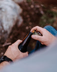A close-up, over-the-shoulder view of a person outdoors by a campfire, holding and opening a dark glass bottle. The person is wearing a light gray shirt and blue jeans, with rocks and earthy ground in the blurred background.