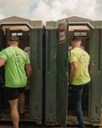 Two men exiting a portable toilet in an outdoor setting with trees and sky. One wearing neon yellow, one wearing neon green Suck less shirts.
