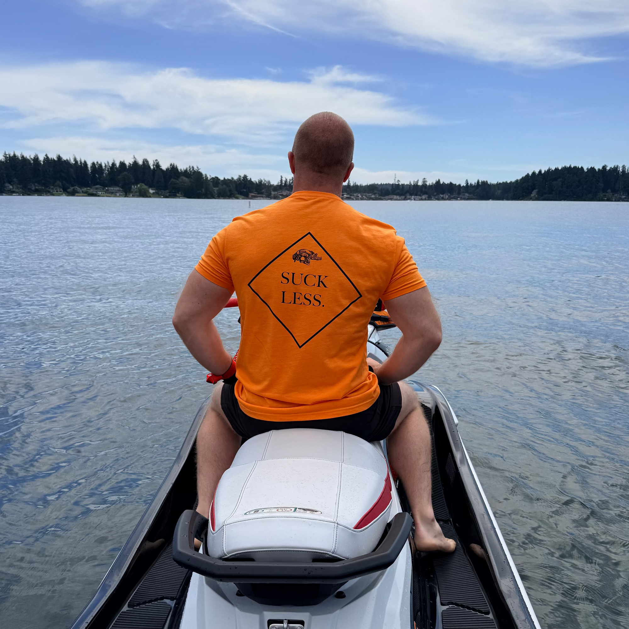 Person sitting on a jet ski wearing an orange shirt with text, looking out over water.