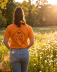 Person wearing an orange shirt with text standing in a field at sunset