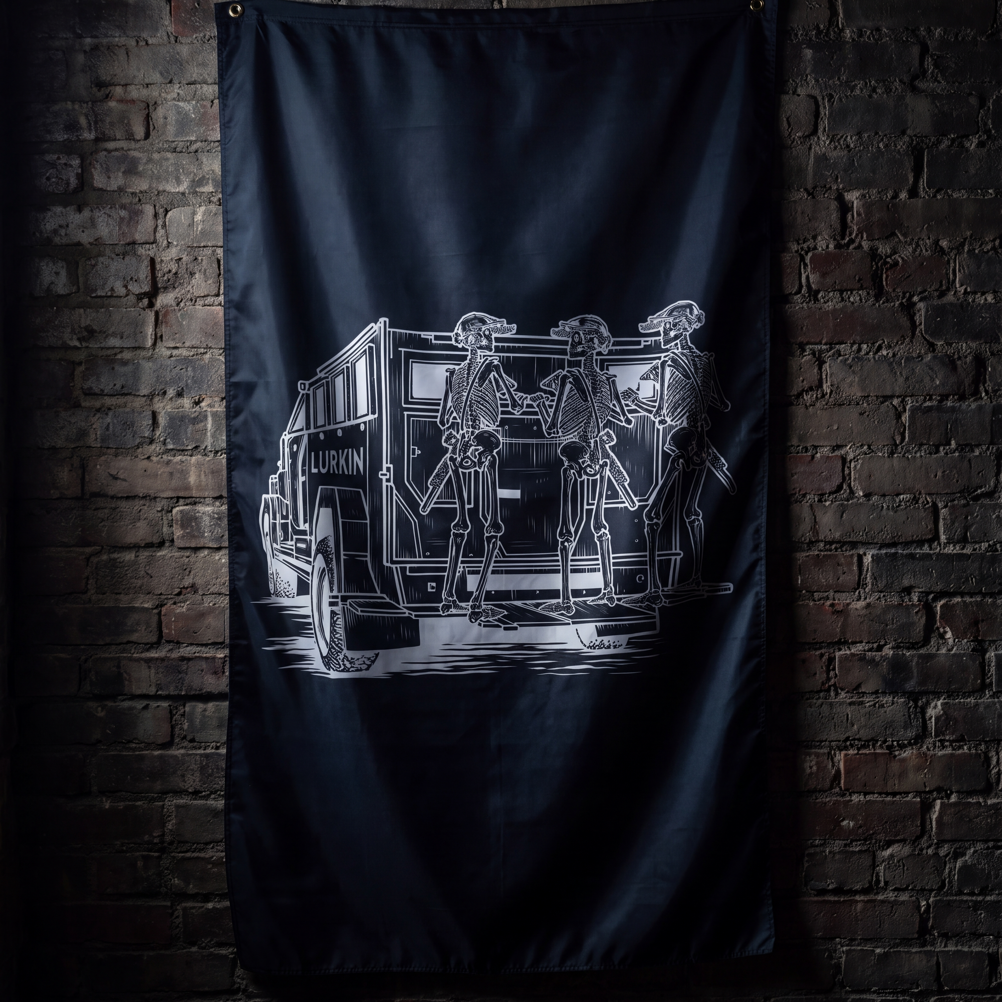 A black flag featuring a white graphic illustration of skeleton figures standing beside a vintage police vehicle, hanging against a dark brick wall.