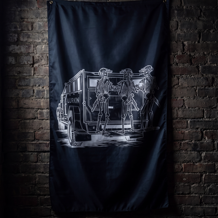 A black flag featuring a white graphic illustration of skeleton figures standing beside a vintage police vehicle, hanging against a dark brick wall.
