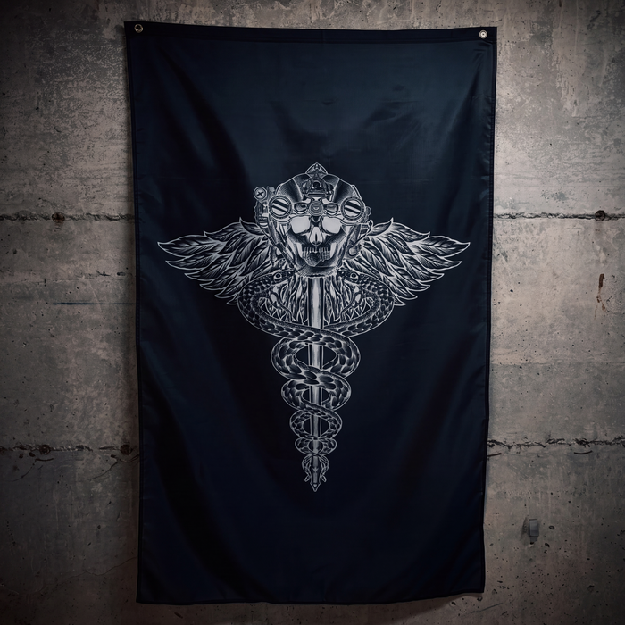 A black flag hanging on a textured concrete wall, featuring a large silver metallic graphic of a winged caduceus skull in the center.