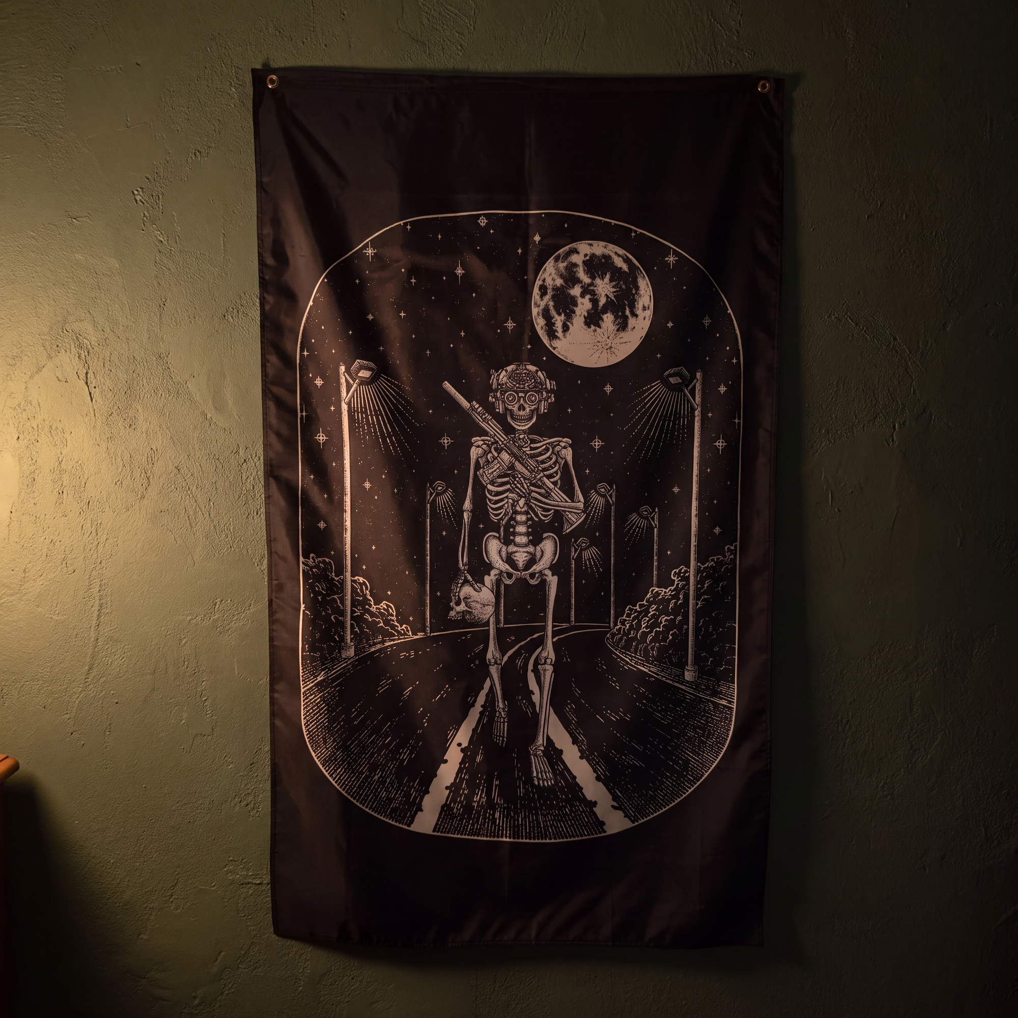 A dark gothic wall flag featuring a skeleton walking down a road under a full moon, hung on a dark textured wall.