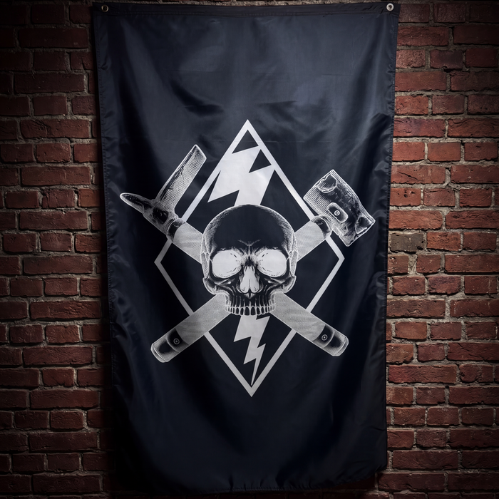 A black flag featuring a white skull and crossed tools graphic, hanging against a red-brown brick wall.