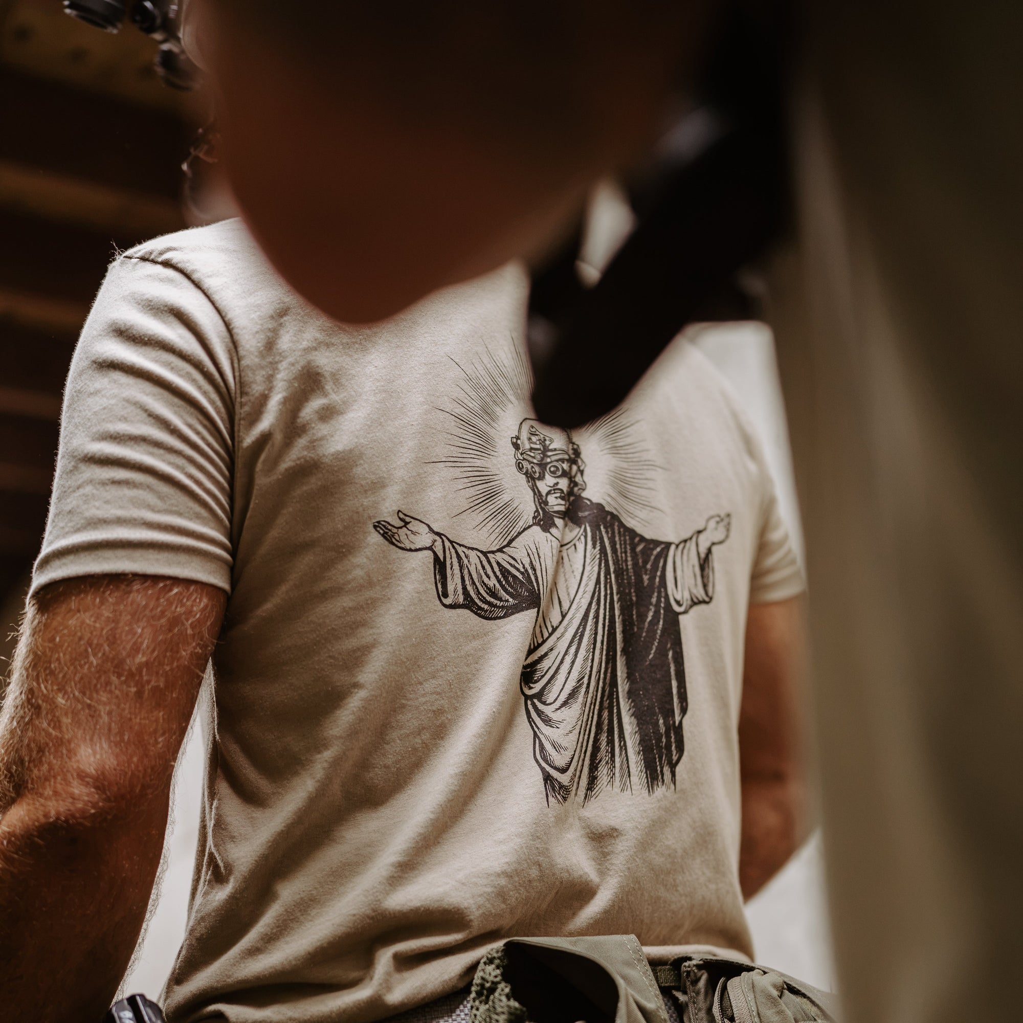 A close-up lifestyle shot of a person wearing a beige t-shirt with a black graphic of Jesus Christ on the back wearing night vision. 