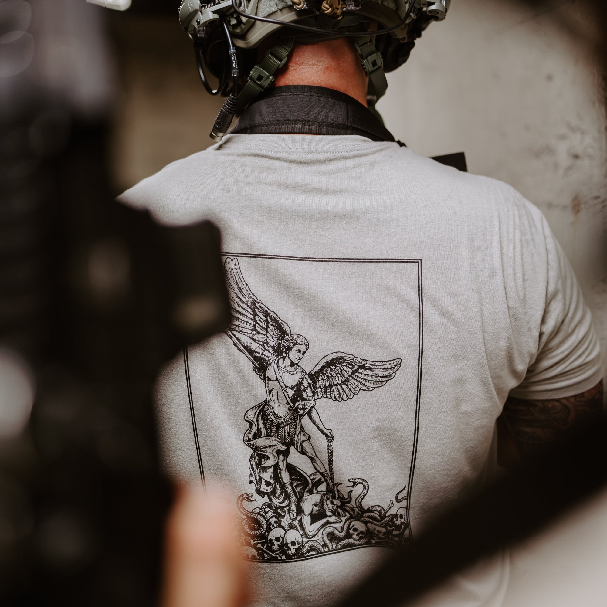 A man viewed from behind wearing a tactical helmet and a light gray t-shirt featuring a large black graphic of an angel on the back. 