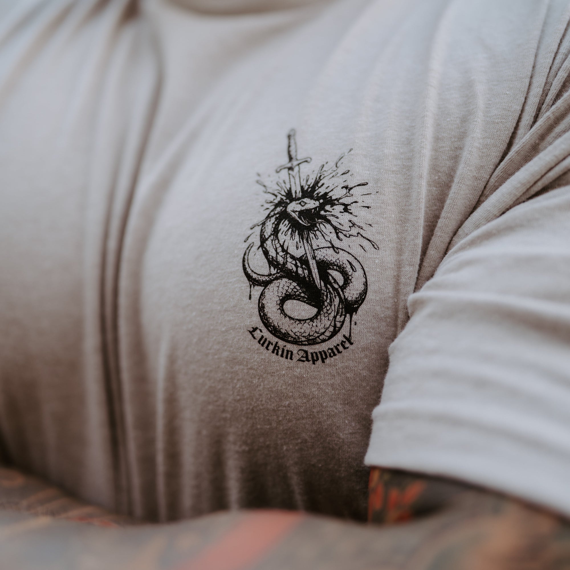 Close-up of a person wearing a light tan t-shirt with a black graphic of a coiled snake and the text 'Lurkin Apparel' printed on the chest.