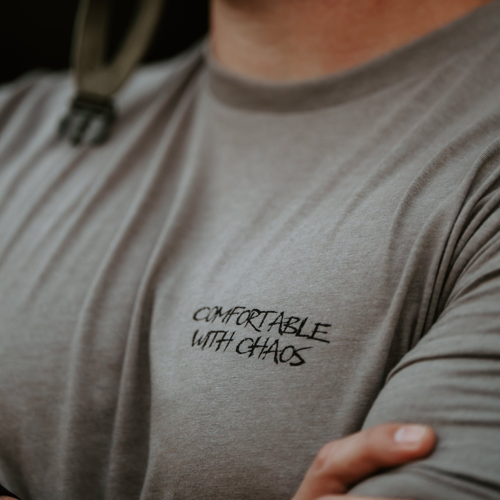 A close-up lifestyle shot of a man wearing a gray crewneck t-shirt with the black printed text 'COMFORTABLE WITH CHAOS' on the left chest. He is also wearing a black tactical headset.