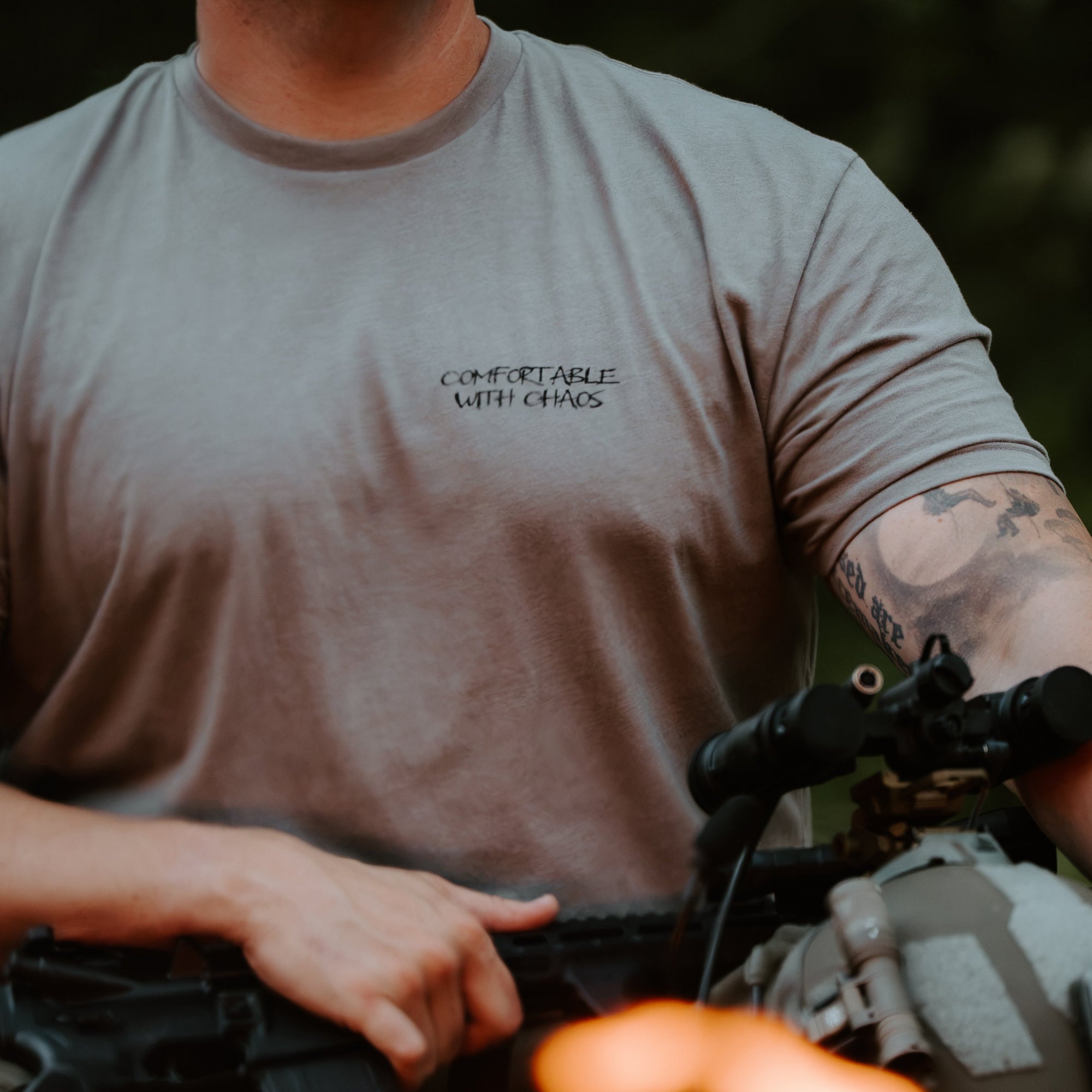 A man wearing a gray short-sleeve t-shirt with the text 'COMFORTABLE WITH CHAOS' printed on the left chest, holding tactical gear in an outdoor setting with blurred flames in the foreground.
