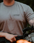 A man wearing a gray short-sleeve t-shirt with the text 'COMFORTABLE WITH CHAOS' printed on the left chest, holding tactical gear in an outdoor setting with blurred flames in the foreground.