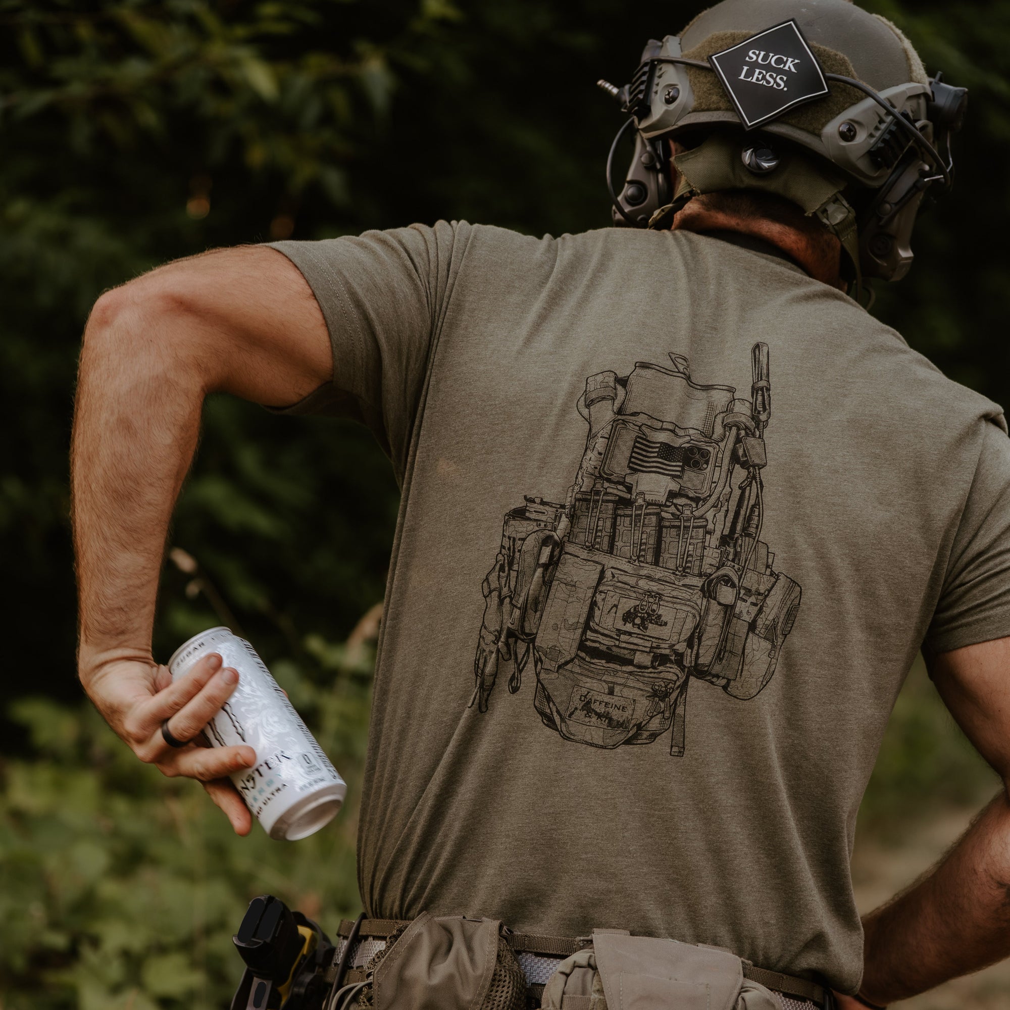 A man wearing a tactical helmet and olive green t-shirt, viewed from behind. The back of the shirt features a detailed black line-art graphic of a tactical vest.