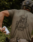 A man wearing a tactical helmet and olive green t-shirt, viewed from behind. The back of the shirt features a detailed black line-art graphic of a tactical vest.