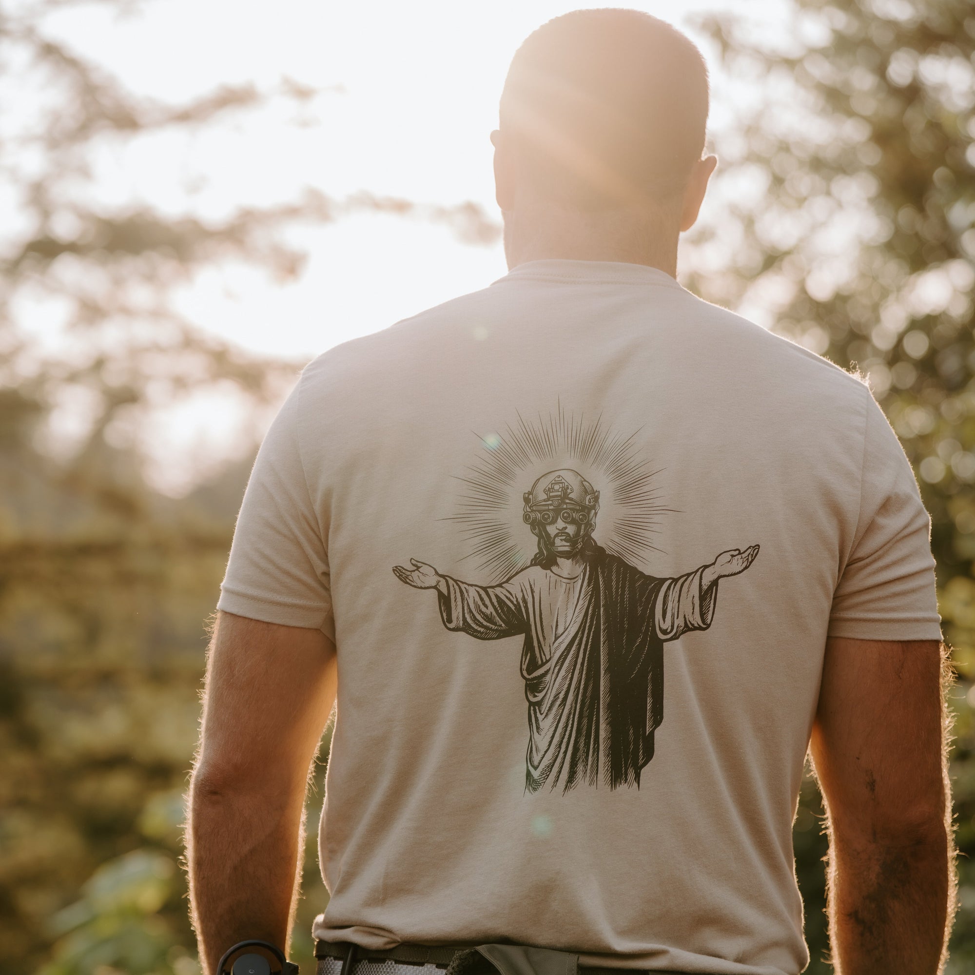 A man viewed from behind wearing a light beige t-shirt with a large black graphic of Jesus Christ on the back, standing outdoors in a sunlit forest setting.