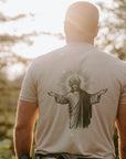 A man viewed from behind wearing a light beige t-shirt with a large black graphic of Jesus Christ on the back, standing outdoors in a sunlit forest setting.