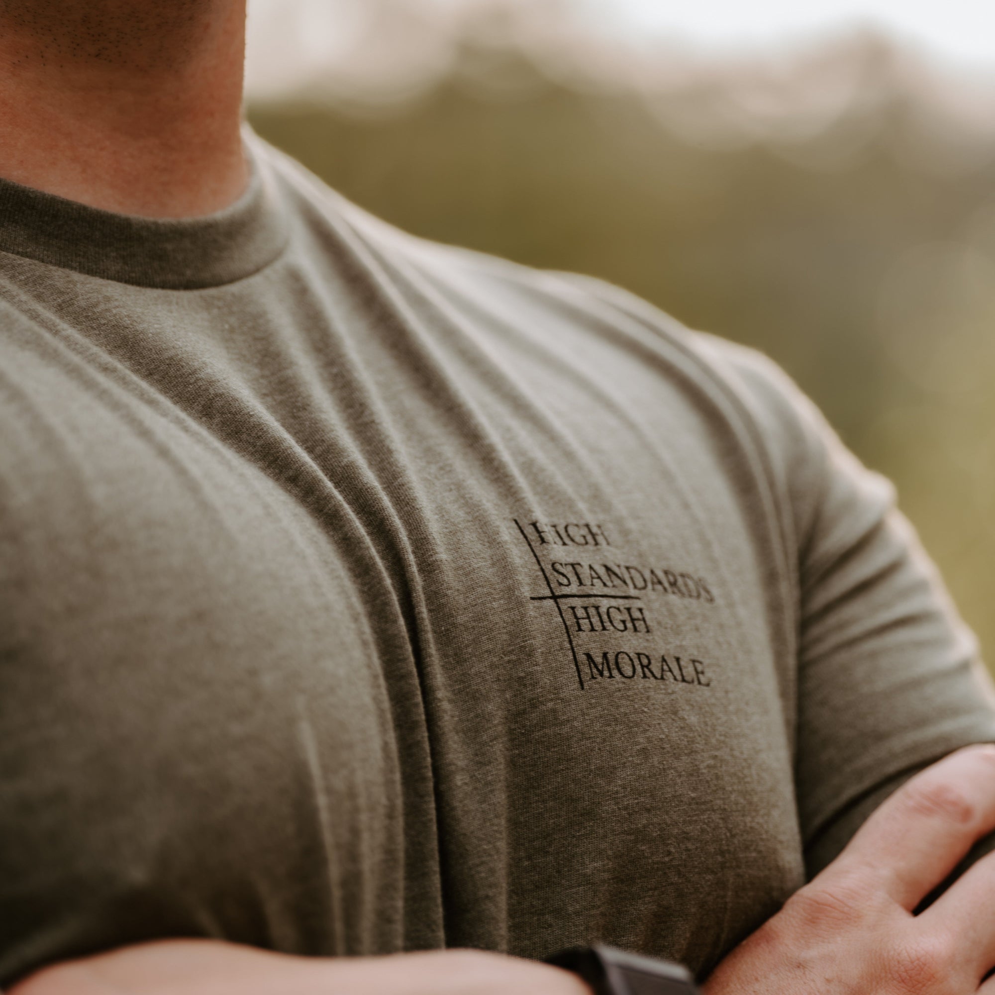 A close-up lifestyle shot of a man wearing a muted olive green t-shirt with black printed text on the left chest that reads 'HIGH STANDARDS HIGH MORALE'. The background is a soft-focus outdoor scene.