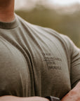 A close-up lifestyle shot of a man wearing a muted olive green t-shirt with black printed text on the left chest that reads 'HIGH STANDARDS HIGH MORALE'. The background is a soft-focus outdoor scene.