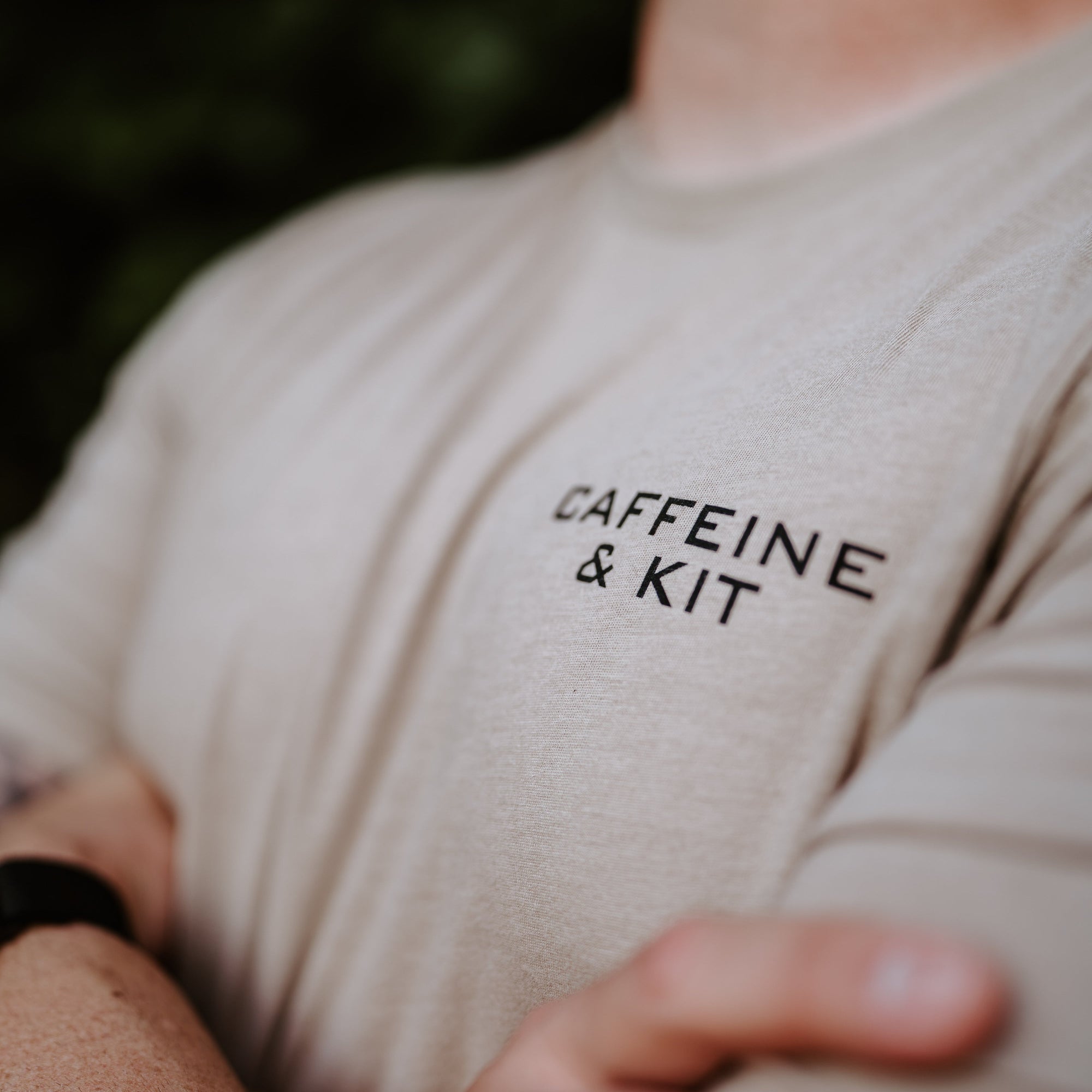A close-up shot of a person wearing a light beige t-shirt with the words 'CAFFEINE & KIT' printed in black on the chest, set against a dark, blurred background.