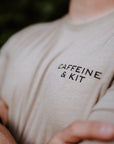 A close-up shot of a person wearing a light beige t-shirt with the words 'CAFFEINE & KIT' printed in black on the chest, set against a dark, blurred background.