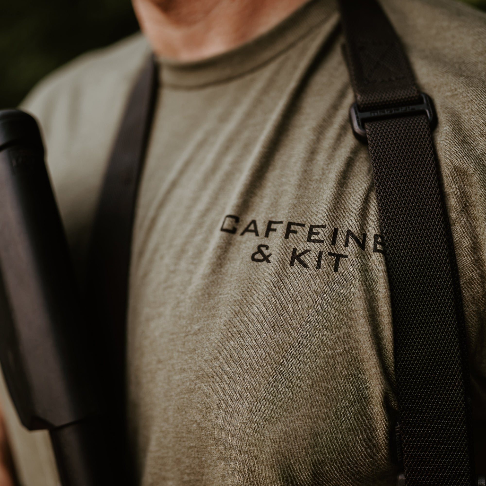 Close-up of a man wearing an olive green t-shirt with 'CAFFEINE & KIT' printed in black on the chest, with black utility straps over his shoulders.