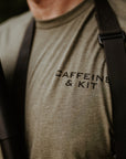 Close-up of a man wearing an olive green t-shirt with 'CAFFEINE & KIT' printed in black on the chest, with black utility straps over his shoulders.