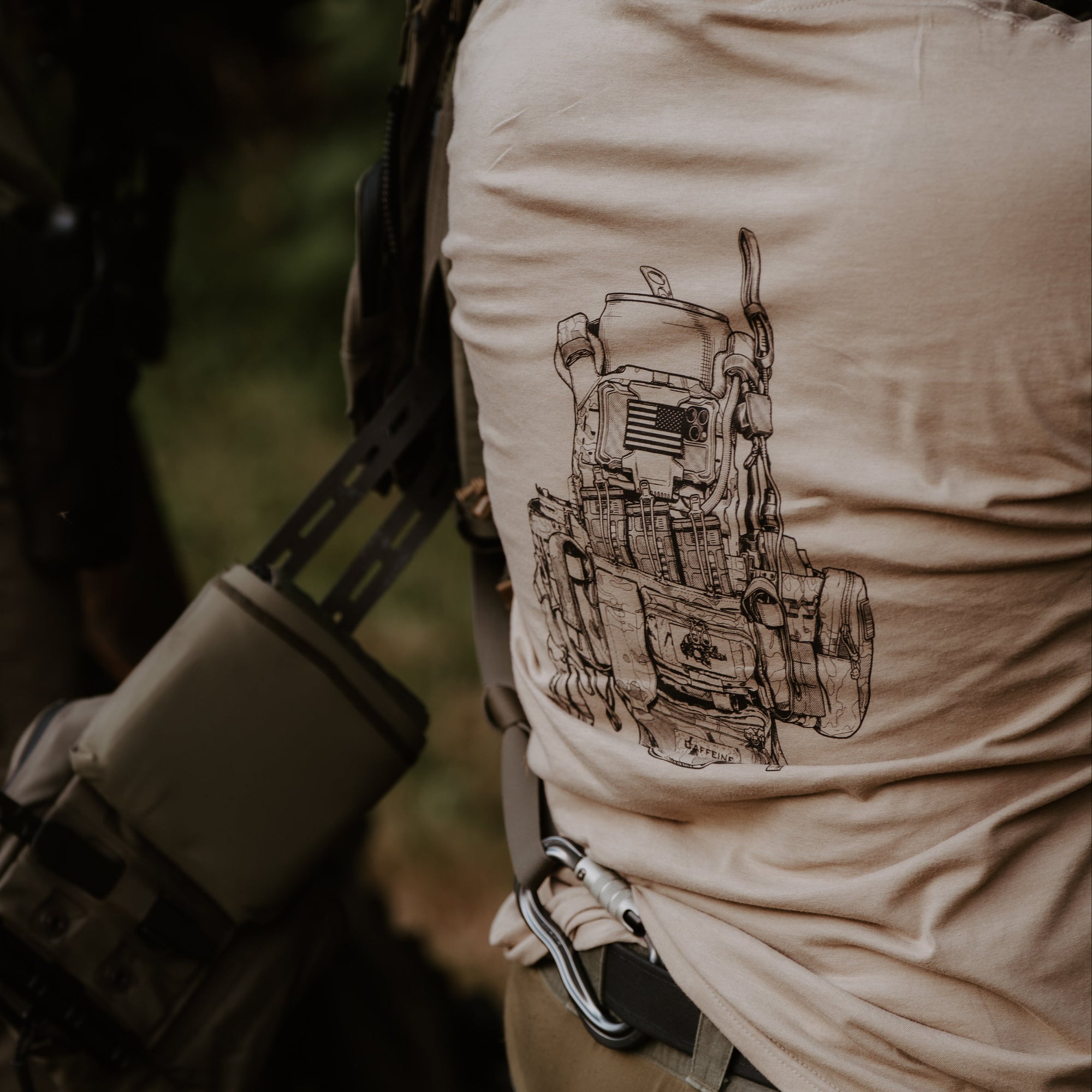 A close-up lifestyle shot of a person outdoors wearing a tan t-shirt with a black line-art graphic of a military-style vest on the back, paired with tactical gear.