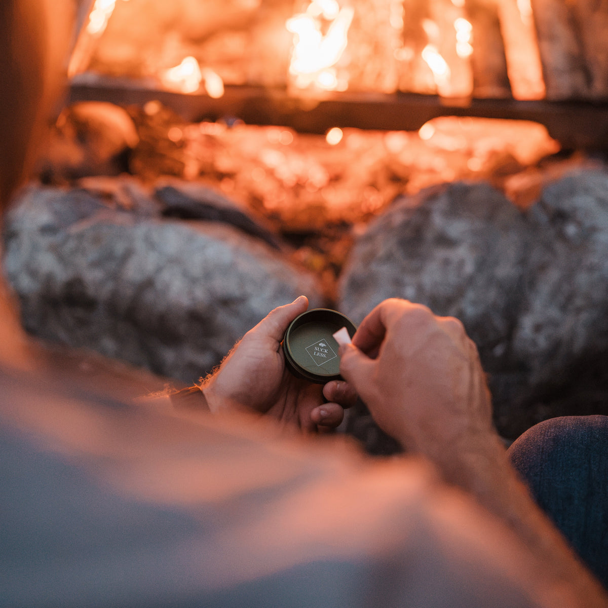 A person sitting by a campfire holds a lurkin pocket container with the glowing fire and rocks in the background.