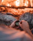 A person sitting by a campfire holds a lurkin pocket container with the glowing fire and rocks in the background.