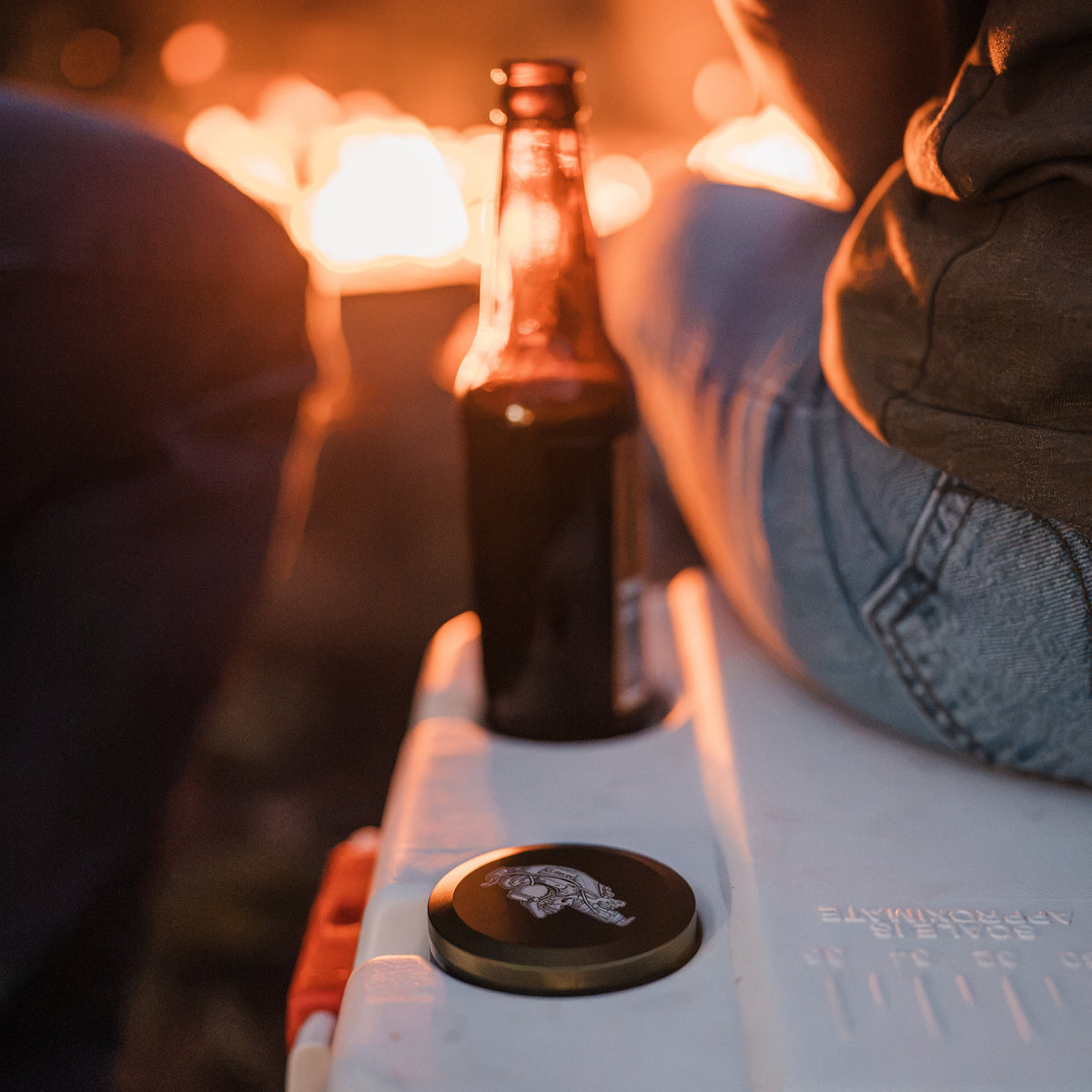 A close-up of the lurkin pocket contianer next to a dark glass container on a cooler by a fire. 