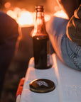 A close-up of the lurkin pocket contianer next to a dark glass container on a cooler by a fire. 