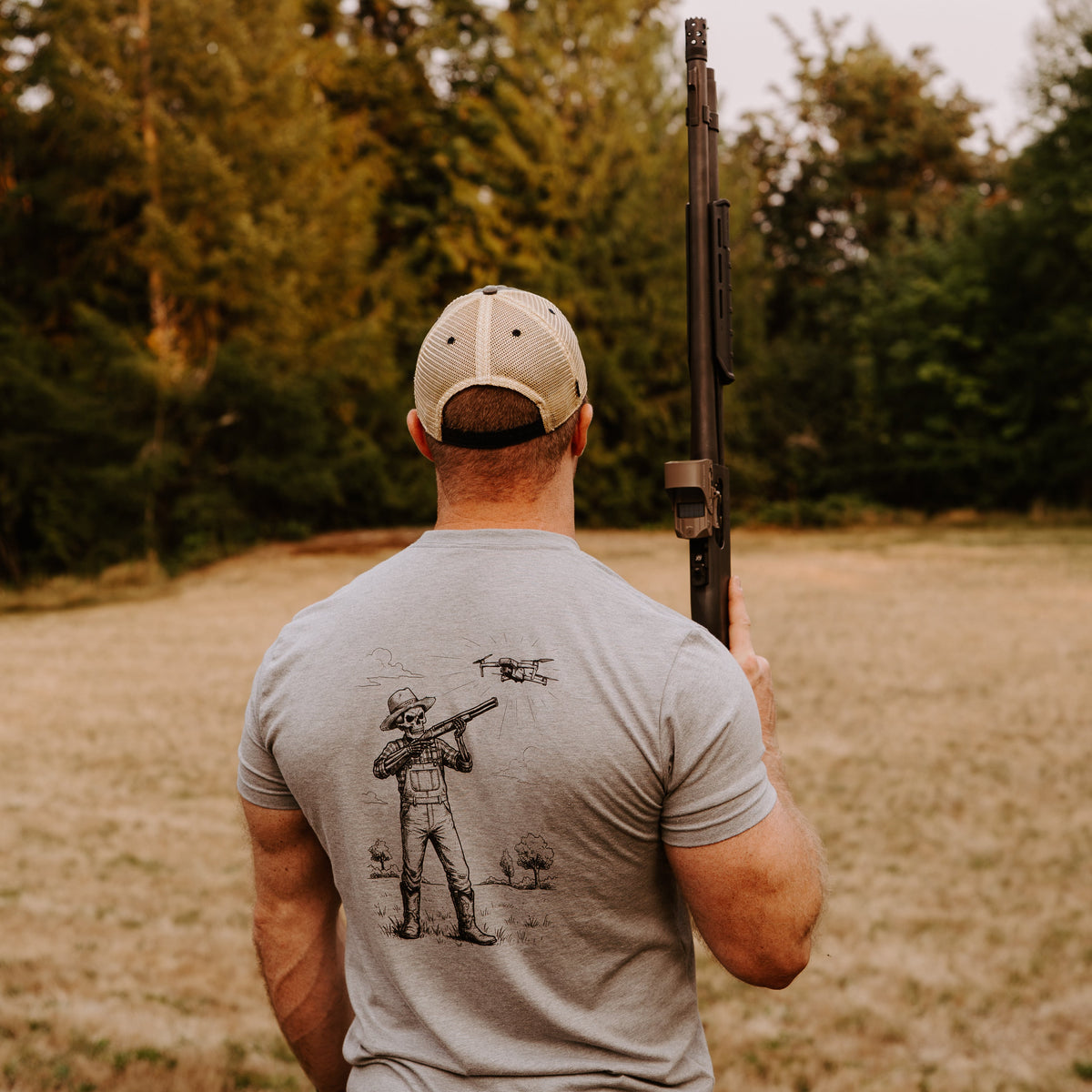 A man viewed from behind wearing a light gray t-shirt with a black Damn Drones graphic on the back, holding a shotgun in an outdoor field with a drone above him. 