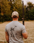 A man viewed from behind wearing a light gray t-shirt with a black Damn Drones graphic on the back, holding a shotgun in an outdoor field with a drone above him. 