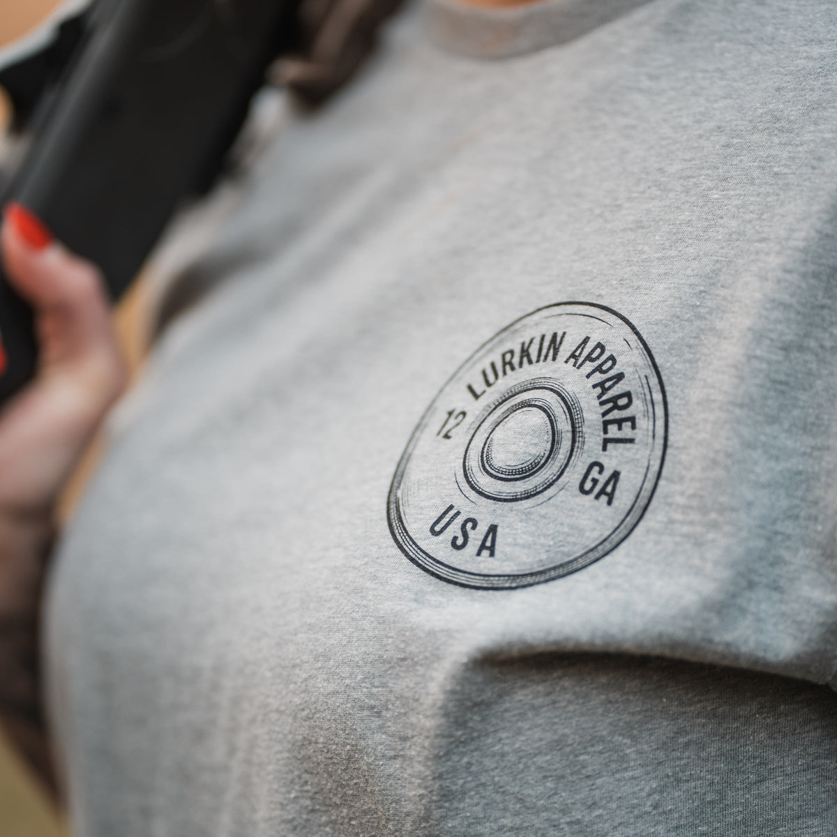 Close-up of a person wearing a heather gray t-shirt with a circular black graphic on the left chest that reads 'LURKIN APPAREL 12 GA USA'.