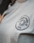 Close-up of a person wearing a heather gray t-shirt with a circular black graphic on the left chest that reads 'LURKIN APPAREL 12 GA USA'.