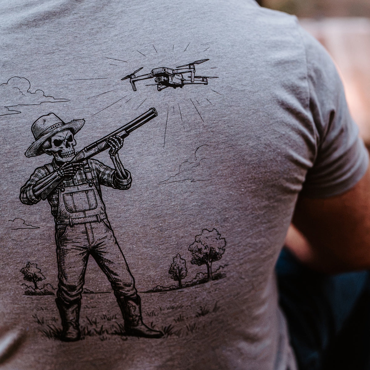 A man wearing a heather gray t-shirt with a black graphic of a skeleton cowboy holding a shotgun under a drone.