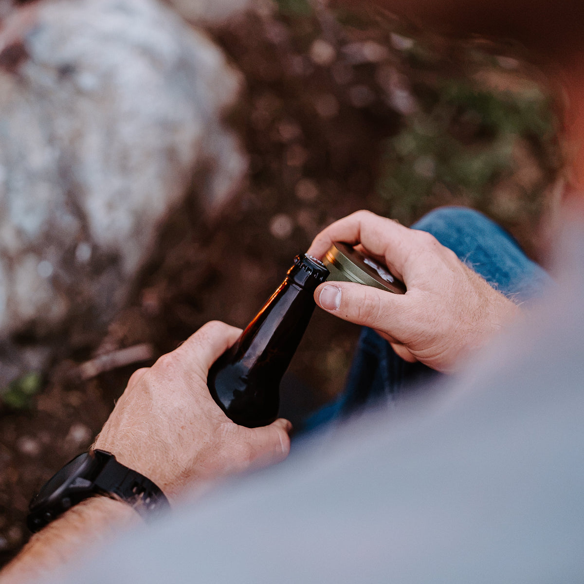 A close-up, over-the-shoulder view of a person outdoors by a campfire, holding and opening a dark glass bottle. The person is wearing a light gray shirt and blue jeans, with rocks and earthy ground in the blurred background.