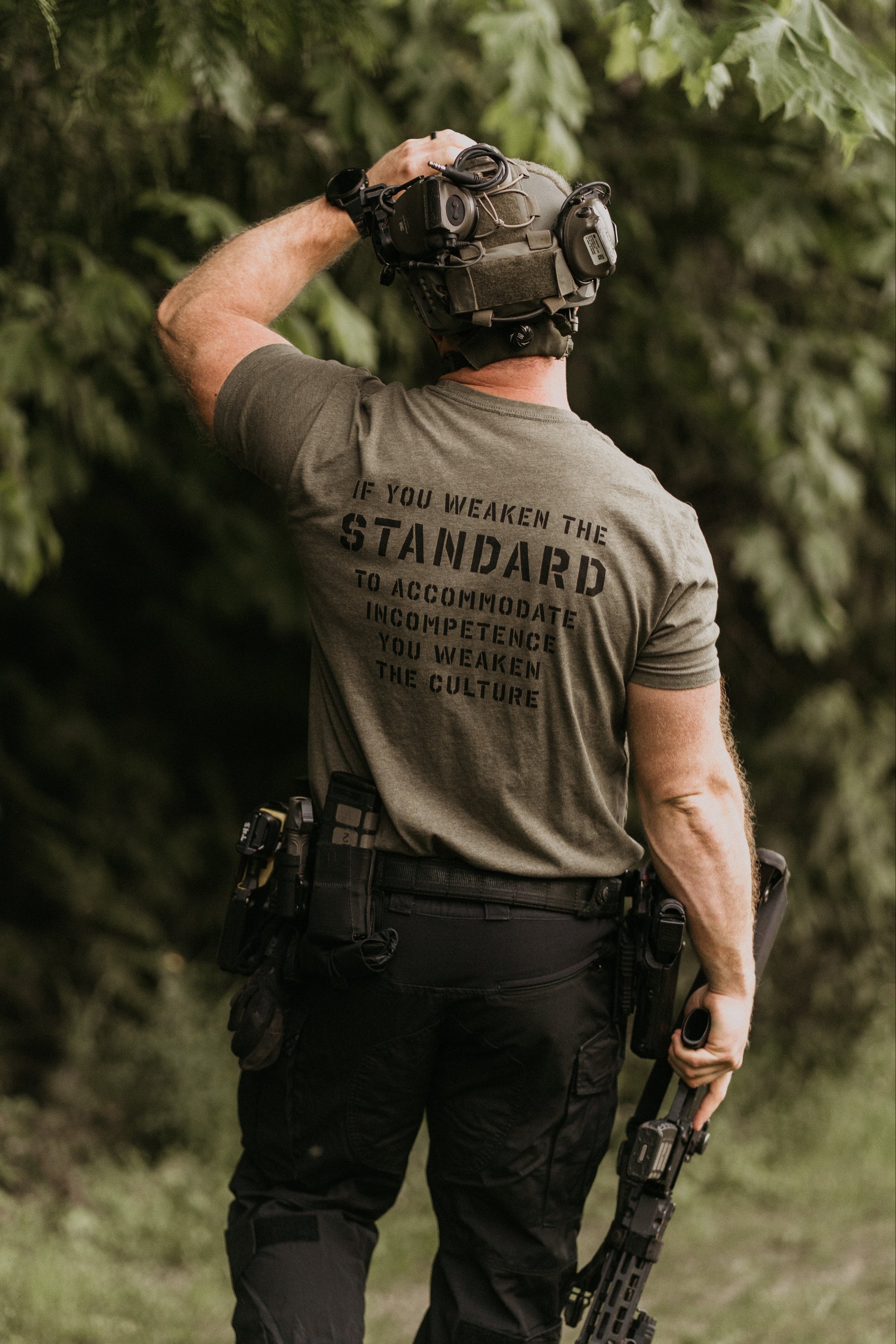 A man wearing tactical gear, including a dark olive green t-shirt with 'STANDARD' printed on the back, black pants, and a helmet, standing in a wooded outdoor environment.