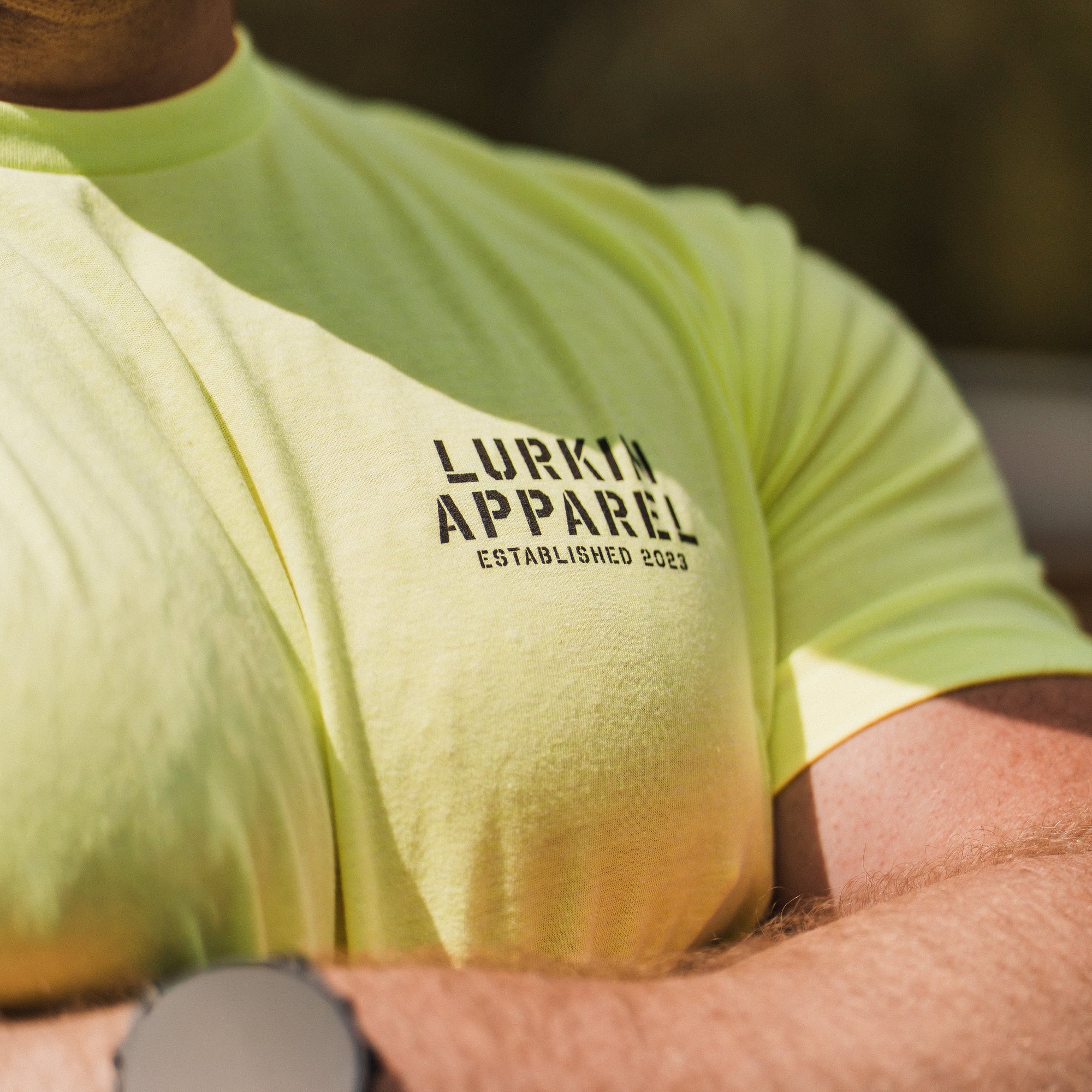 Person wearing a bright yellow t-shirt with 'Lurkin Apparel' branding, outdoors.