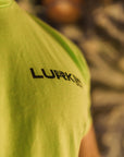 Person wearing a neon green t-shirt with 'Lurkin' logo, blurred background