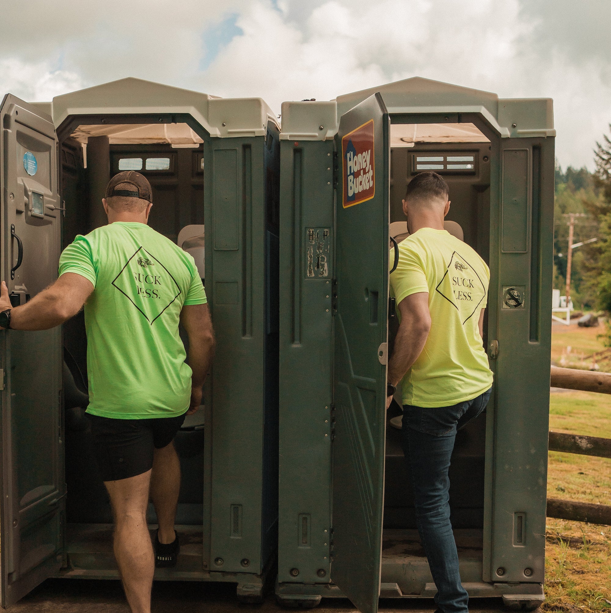 Two men exiting a portable toilet in an outdoor setting with trees and sky. One wearing neon yellow, one wearing neon green Suck less shirts. 
